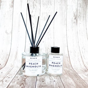 May include: Two glass diffuser bottles with black reeds. The larger bottle contains 150ml of liquid and the smaller bottle contains 70ml. Both bottles have a black cap and a white label that reads "Peach Magnolia" and "All Natural Diffuser".