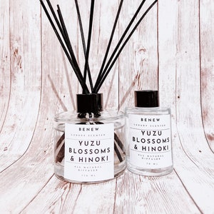 May include: Two glass diffuser bottles with black reeds. The larger bottle has a capacity of 110 ml, and the smaller one is 70 ml. Both bottles have labels that read "Yuzu Blossoms & Hinoki" and "All Natural Diffuser."