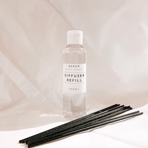 May include: A clear glass bottle with a white label that reads "BENEW LUXURY SCENTED DIFFUSER REFILL 200ML". The bottle is sitting on a white surface next to a bundle of black diffuser sticks.