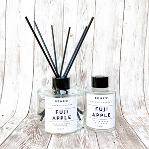 May include: Two glass diffuser bottles with black reeds. The larger bottle is labeled "BENEW LUXURY SCENTED FUJI APPLE ALL NATURAL DIFFUSER 150ml". The smaller bottle is labeled "BENEW LUXURY SCENTED FUJI APPLE ALL NATURAL DIFFUSER 70ml".