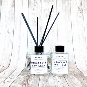 May include: Two clear glass bottles with black lids and black reeds. The bottles are labeled "BENEW LUXURY SCENTED TOBACCO & BAY LEAF ALL NATURAL DIFFUSER 100ML".
