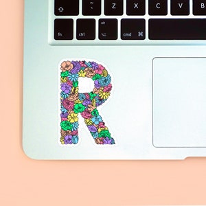 Peach background with the left corner of a MacBook shown on the right with the R floral letter sticker design placed upon it for size reference.