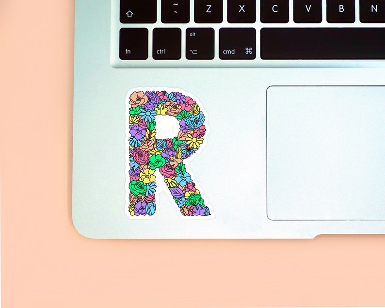 Peach background with the left corner of a MacBook shown on the right with the R floral letter sticker design placed upon it for size reference.