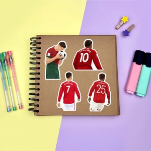 Yellow & purple background with a brown scrapbook in the centre, gel pens & highlighters placed around it. Several stickers featuring Manchester United men’s footballers on the scrapbook page for size reference.