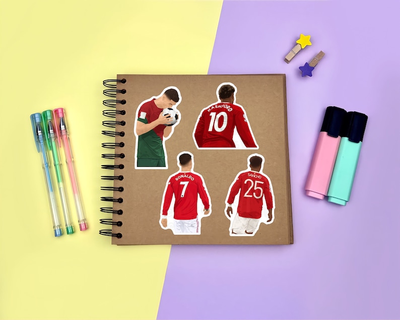 Yellow & purple background with a brown scrapbook in the centre, gel pens & highlighters placed around it. Several stickers featuring Manchester United men’s footballers on the scrapbook page for size reference.