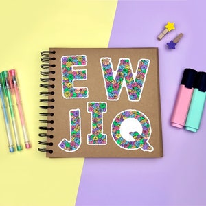 Yellow & purple background with a brown scrapbook in the centre, gel pens & highlighters placed around it. The E, W, J, I & Q floral letter sticker designs placed across the scrapbook page for size reference.