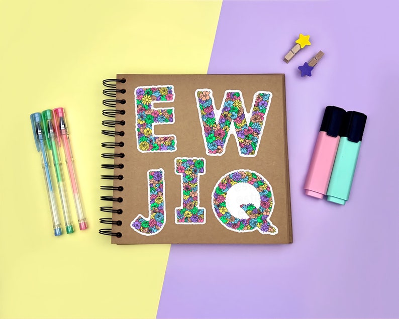 Yellow & purple background with a brown scrapbook in the centre, gel pens & highlighters placed around it. The E, W, J, I & Q floral letter sticker designs placed across the scrapbook page for size reference.