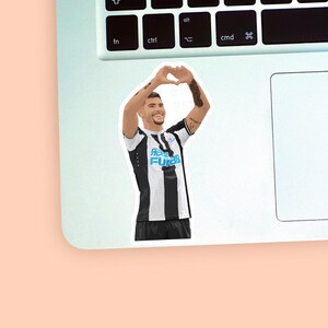 Left corner of a MacBook placed on a peach background with the Bruno Guimaraes sticker design placed on it for size reference.