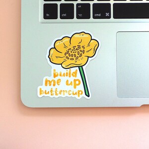 Left corner of a MacBook on a peach background with the Build me up buttercup vinyl sticker placed on it for size reference.