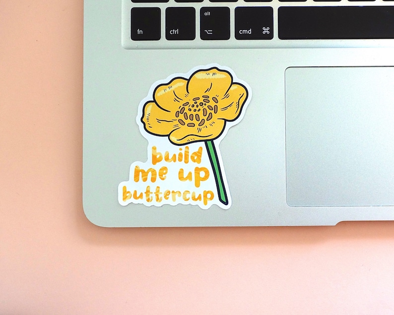 Left corner of a MacBook on a peach background with the Build me up buttercup vinyl sticker placed on it for size reference.