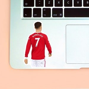 Peach background with the left corner of a MacBook & the sticker design of Ronaldo wearing a red Manchester United kit placed on it for size reference. The design is drawn from the rear to highlight the number 7 & Ronaldo on the back of his shirt.