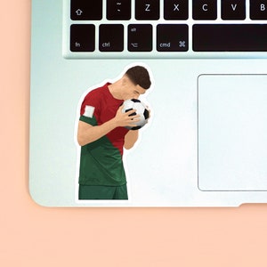 Peach background with the left corner of a MacBook & the second sticker design of Ronaldo wearing a red & green Portugal kit placed on it for size reference. The design is drawn from the side showing Ronaldo holding a ball in his hands & kissing it.