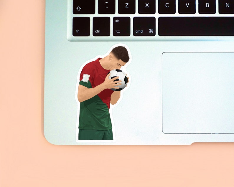 Peach background with the left corner of a MacBook & the second sticker design of Ronaldo wearing a red & green Portugal kit placed on it for size reference. The design is drawn from the side showing Ronaldo holding a ball in his hands & kissing it.