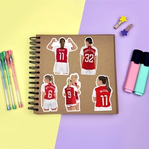 Yellow & purple background with a brown scrapbook in the centre, gel pens & highlighters placed around it. Several stickers featuring Arsenal women’s footballers on the scrapbook page for size reference.