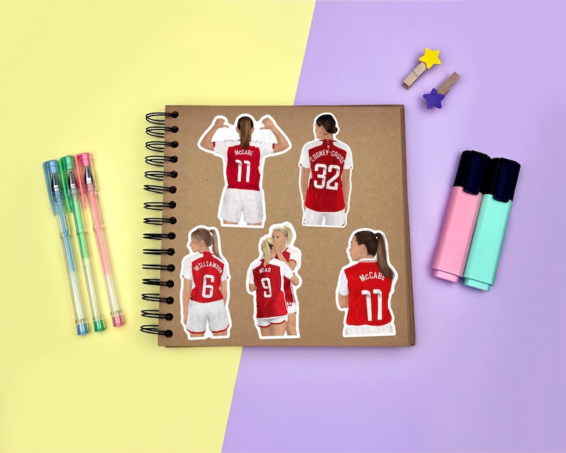 Yellow & purple background with a brown scrapbook in the centre, gel pens & highlighters placed around it. Several stickers featuring Arsenal women’s footballers on the scrapbook page for size reference.