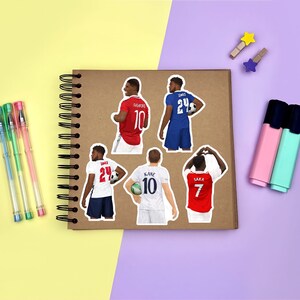 Yellow & purple background with a brown scrapbook in the centre, gel pens & highlighters placed around it. Several sticker designs of men’s footballers placed across the scrapbook page for size reference.