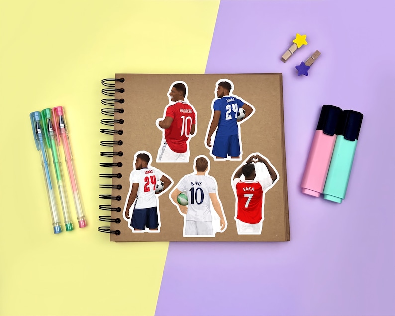 Yellow & purple background with a brown scrapbook in the centre, gel pens & highlighters placed around it. Several sticker designs of men’s footballers placed across the scrapbook page for size reference.