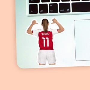 Left corner of a MacBook placed on a peach background with the second Katie McCabe sticker design placed on it for size reference.