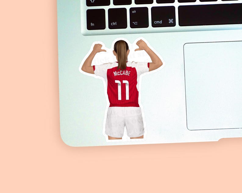 Left corner of a MacBook placed on a peach background with the second Katie McCabe sticker design placed on it for size reference.
