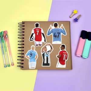 Yellow & purple background with a brown scrapbook in the centre, gel pens & highlighters placed around it. Several stickers featuring men’s footballers on the scrapbook page for size reference.