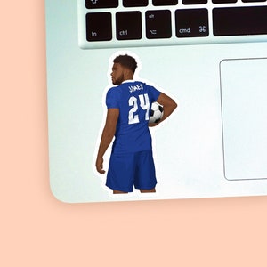Left corner of a MacBook on a peach background with a vinyl sticker of footballer Reece James wearing the royal blue Chelsea football kit placed on it for size reference.