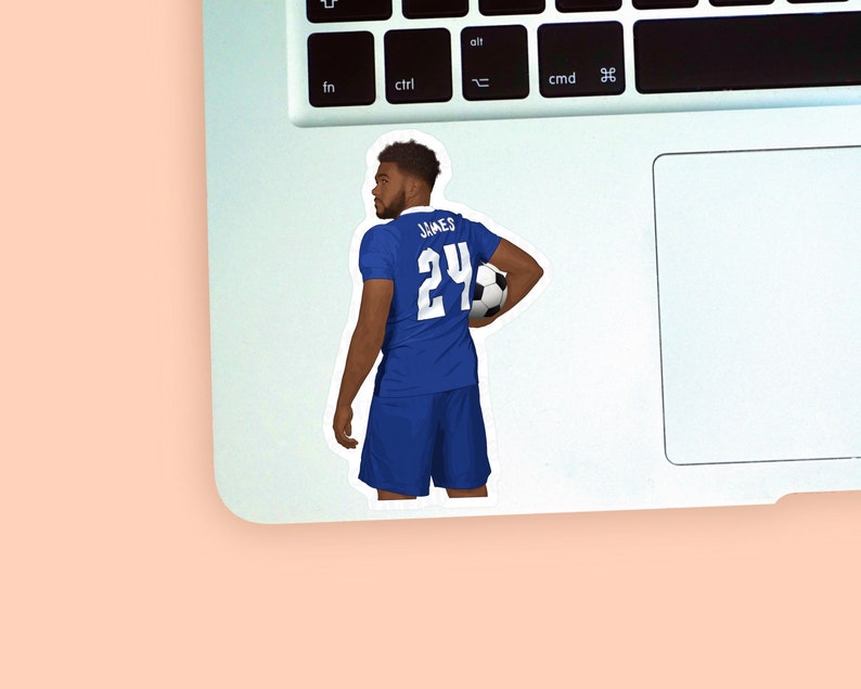 Left corner of a MacBook on a peach background with a vinyl sticker of footballer Reece James wearing the royal blue Chelsea football kit placed on it for size reference.