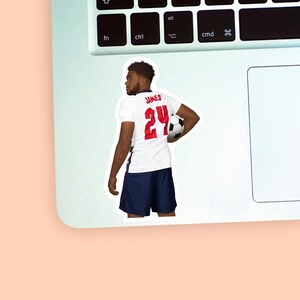 Left corner of a MacBook on a peach background with a vinyl sticker of footballer Reece James wearing the white & blue england football kit placed on it for size reference.