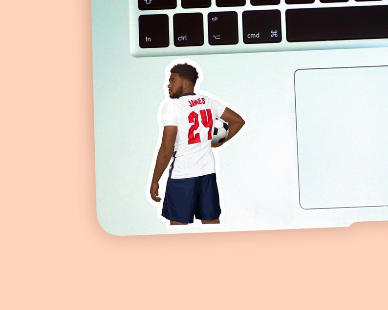 Left corner of a MacBook on a peach background with a vinyl sticker of footballer Reece James wearing the white & blue england football kit placed on it for size reference.