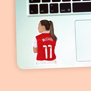 Left corner of a MacBook placed on a peach background with the first Katie McCabe sticker design placed on it for size reference.