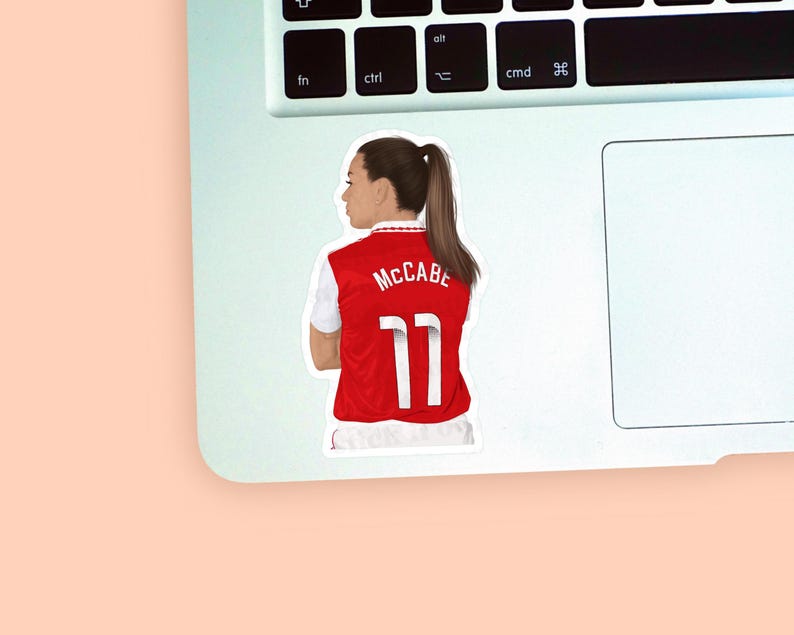 Left corner of a MacBook placed on a peach background with the first Katie McCabe sticker design placed on it for size reference.