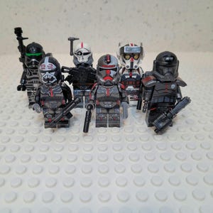 May include: A collection of six plastic toy soldier minifigures featuring detailed armor in shades of black, gray, white, and red. Each figure is equipped with various molded plastic weapons and distinct helmet designs, standing in a group formation.
