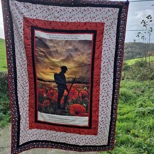 May include: A patchwork quilt with a central image of a soldier in a poppy field at sunset. The image is framed by red and black borders, and a white background with red floral accents. Text is visible on the panel.
