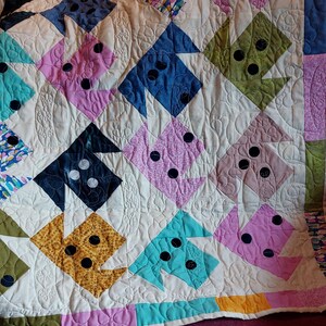 Puede incluir: Una colcha blanca con un diseño de patchwork que presenta varios colores y patrones, incluyendo rosa, azul, verde, amarillo y negro. La colcha está doblada y drapeada sobre un sofá burdeos.