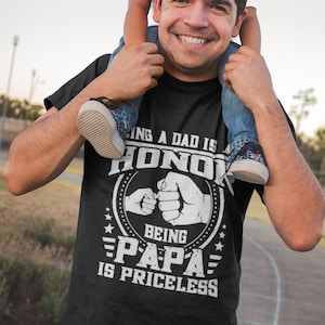 K&ouml;nnte beinhalten: Ein schwarzes T-Shirt mit einem wei&szlig;en Aufdruck, der "Ein Vater zu sein ist eine Ehre, Papa zu sein ist unbezahlbar" lautet. Die Grafik zeigt auch zwei sich sto&szlig;ende F&auml;uste. Ein Mann tr&auml;gt das Shirt und hat ein Kind auf seinen Schultern.