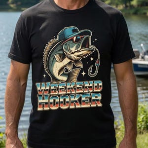 May include: Black t-shirt with a graphic of a fish wearing sunglasses and a hat, holding a fishing hook. The text "WEEKEND HOOKER" is below the fish in a retro font. A man is wearing the shirt in front of a lake.