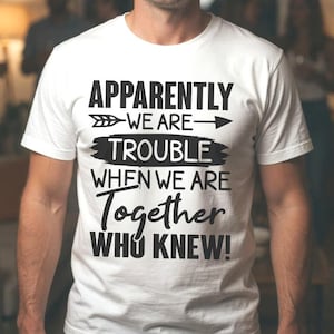 Può includere: T-shirt bianca con la scritta "Apparently We Are Trouble When We Are Together Who Knew!" in nero. La maglietta è indossata da una persona sorridente in una stanza con altre persone sullo sfondo.