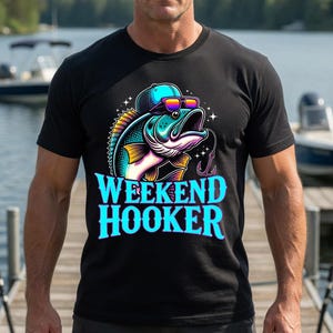 May include: Black t-shirt featuring a colorful graphic of a fish wearing sunglasses and a hat, with the text "WEEKEND HOOKER" in blue and white. The image is set against a lake backdrop.