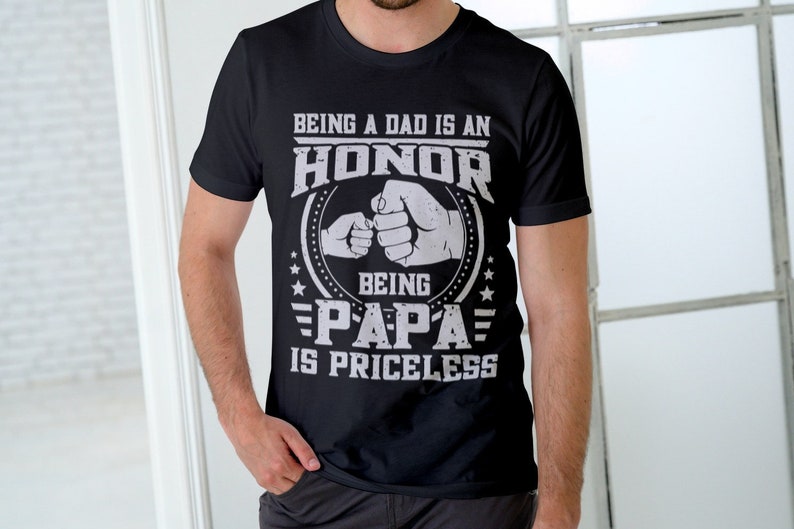 K&ouml;nnte beinhalten: Schwarzes T-Shirt mit einer wei&szlig;en Grafik, auf der steht "Ein Vater zu sein ist eine Ehre, Papa zu sein ist unbezahlbar". Die Grafik zeigt zwei aufeinanderprallende F&auml;uste, eine gr&ouml;&szlig;ere und eine kleinere, mit Sternen und Streifen um die F&auml;uste.