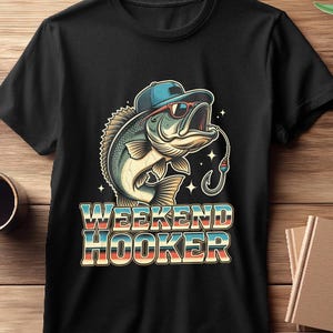 May include: Black t-shirt featuring a cartoon bass wearing sunglasses and a cap, with a fishing hook in its mouth. The text "WEEKEND HOOKER" is displayed in a retro font, with blue, red, and white colors. The shirt is on a wooden surface.