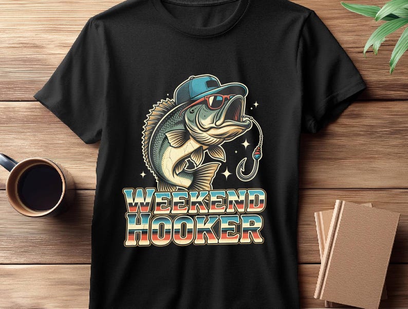 May include: Black t-shirt featuring a cartoon bass wearing sunglasses and a cap, with a fishing hook in its mouth. The text "WEEKEND HOOKER" is displayed in a retro font, with blue, red, and white colors. The shirt is on a wooden surface.
