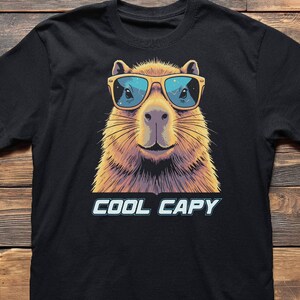 May include: A black short-sleeved t-shirt featuring a graphic illustration of a capybara wearing yellow-framed sunglasses with blue lenses. Below the capybara, the text COOL CAPY is printed in a bold, light blue font with a dark outline.