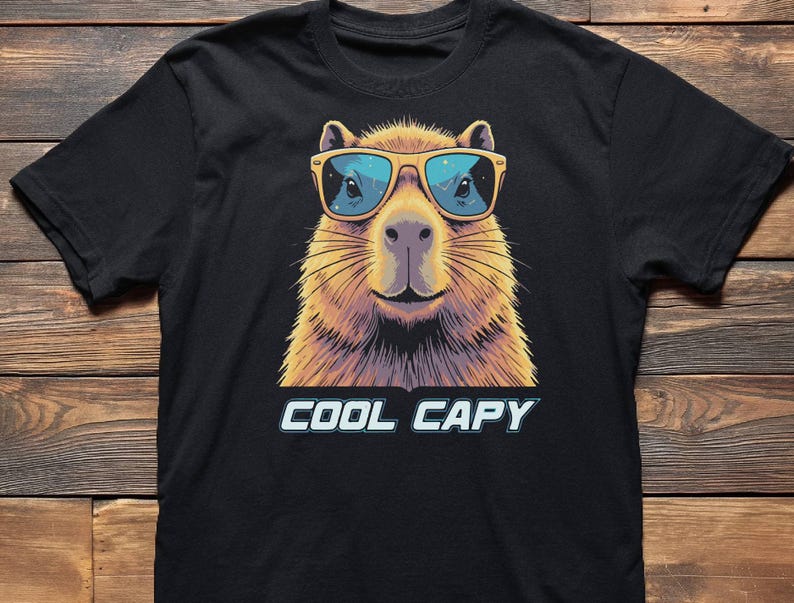 May include: A black short-sleeved t-shirt featuring a graphic illustration of a capybara wearing yellow-framed sunglasses with blue lenses. Below the capybara, the text COOL CAPY is printed in a bold, light blue font with a dark outline.