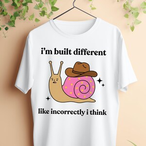 May include: White t-shirt with the text "I'm built different like incorrectly I think." The graphic features a cartoon snail wearing a brown cowboy hat and a pink shell with yellow stars. Two black sparkles are on either side of the snail.