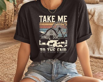 May include: Black t-shirt with a graphic featuring a barn, Ferris wheel, and farm animals. The text "TAKE ME TO THE FAIR" is printed above the scene in white block letters. The shirt is worn by a person.