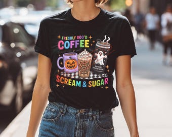 May include: A black t-shirt featuring a colorful graphic with a purple pumpkin mug, a spiderweb-patterned coffee cup, and a ghost-themed tumbler. The text reads FRESHLY BOO'D COFFEE and SCREAM & SUGAR, surrounded by small stars and a checkered base.