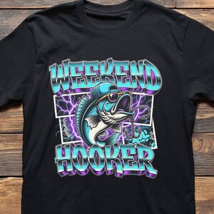 May include: Black t-shirt featuring a cartoon fish wearing sunglasses and a cap, with the words "WEEKEND HOOKER" in a blue and purple gradient font. The design includes a fishing hook and lightning bolts.