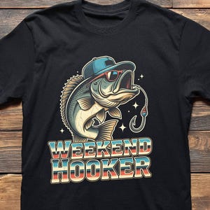 May include: Black t-shirt featuring a cartoon bass wearing a blue cap and sunglasses, with a fishing hook in its mouth. The text "WEEKEND HOOKER" is displayed in a retro font, with blue, red, and cream colors.