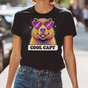 May include: A black short-sleeve t-shirt featuring a graphic of a capybara wearing pink heart-shaped sunglasses with a galaxy pattern. The capybara has a smiling expression with visible teeth. Below the graphic, the text COOL CAPY is printed.