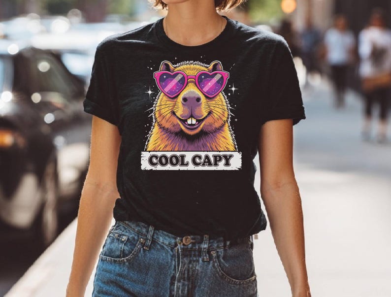 May include: A black short-sleeve t-shirt featuring a graphic of a capybara wearing pink heart-shaped sunglasses with a galaxy pattern. The capybara has a smiling expression with visible teeth. Below the graphic, the text COOL CAPY is printed.