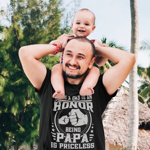 K&ouml;nnte beinhalten: Ein schwarzes T-Shirt mit einem wei&szlig;en Grafikdesign, auf dem "Ein Vater zu sein ist eine Ehre, Papa zu sein ist unbezahlbar" steht. Die Grafik zeigt auch zwei sich sto&szlig;ende F&auml;uste. Ein Mann tr&auml;gt das Shirt und tr&auml;gt ein kleines Kind auf seinen Schultern.